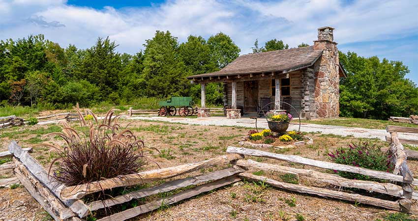 Historic log cabin atop Baird Mountain with wood fence and spring flowers in foreground, trees and blue sky behind it.